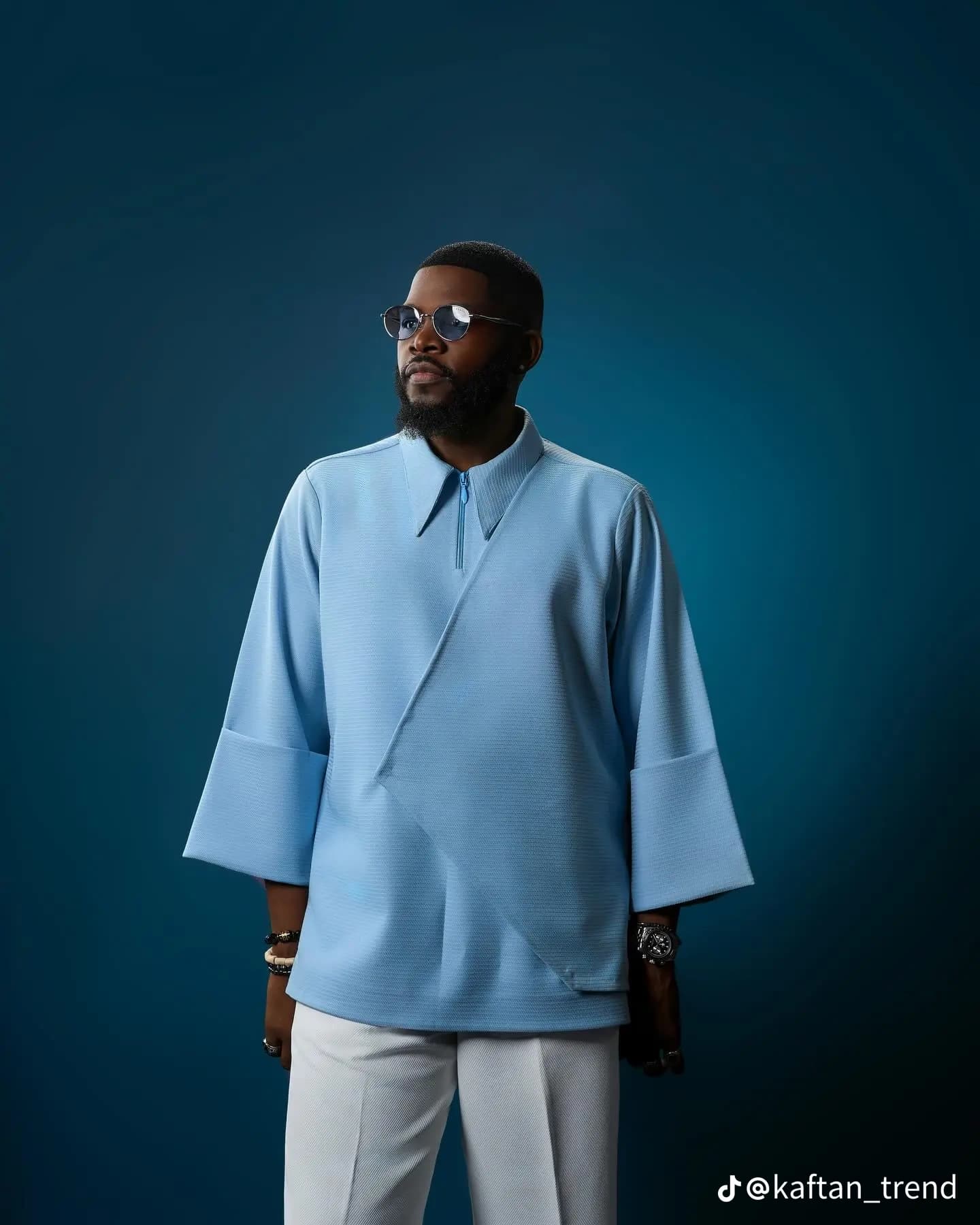 Bearded man wearing a light blue textured tunic and white trousers against a blue background.