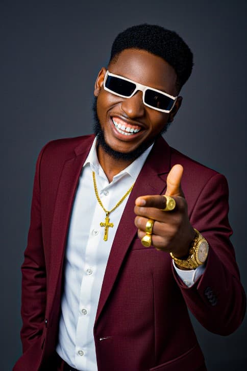Smiling Black man in a maroon suit and sunglasses, pointing forward while wearing gold jewelry.