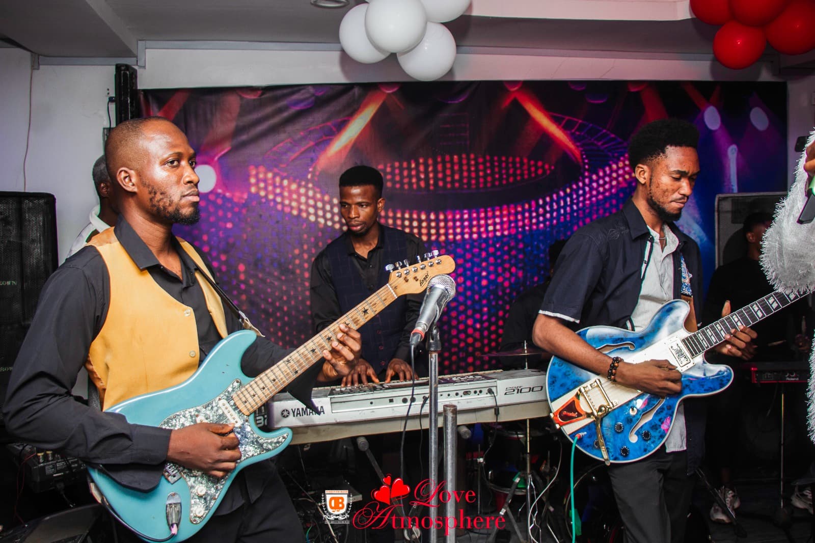 Two guitarists and a keyboardist perform live on stage with a vibrant backdrop.