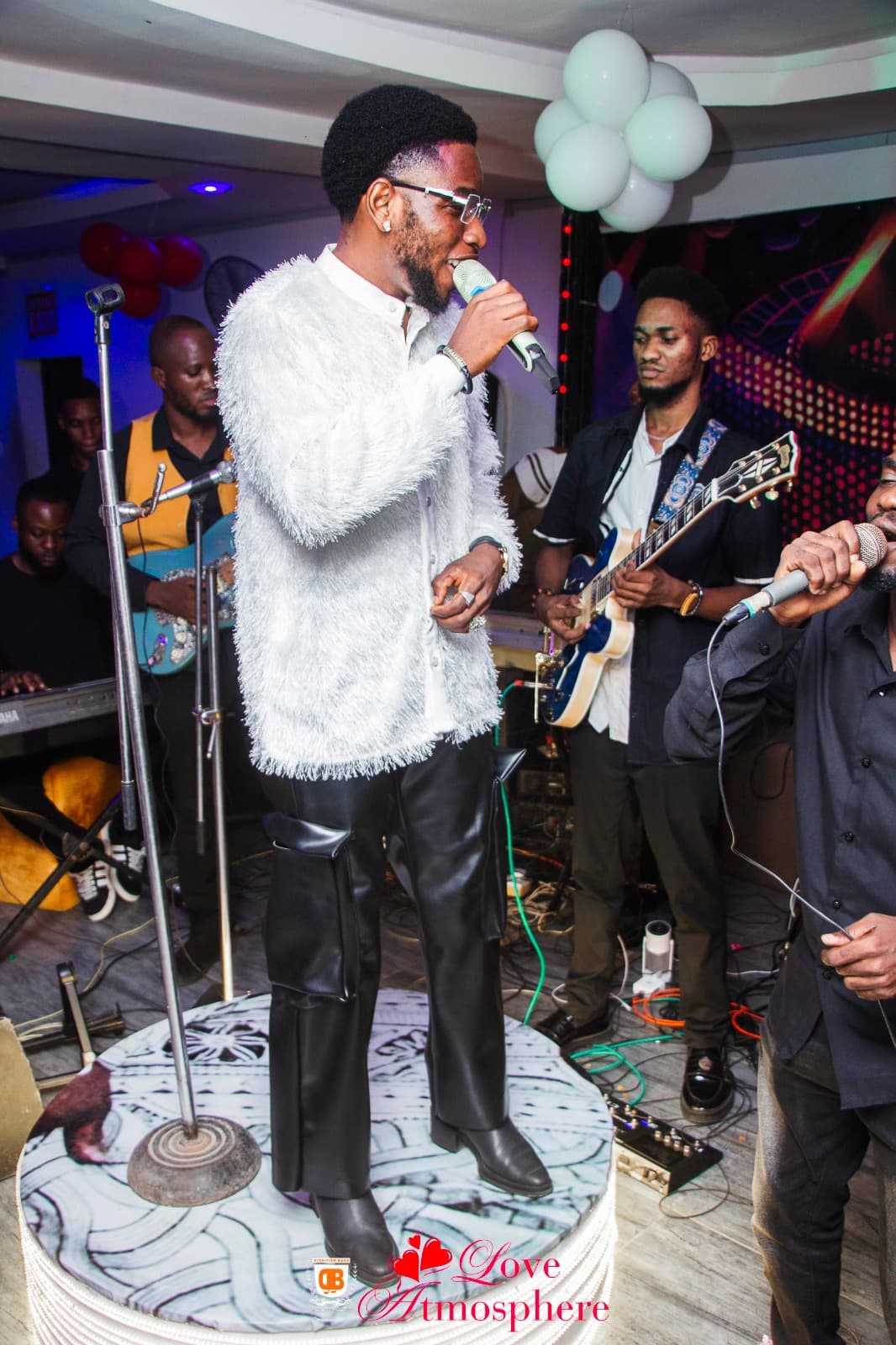 Singer in white shaggy jacket and black leather pants performs on stage with a band.