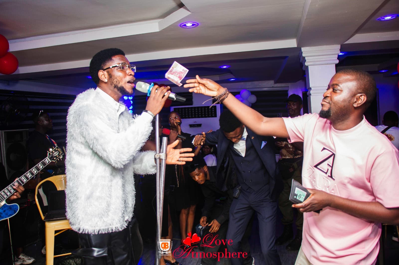 Singer in a white fuzzy jacket performs while a man in pink tosses money.