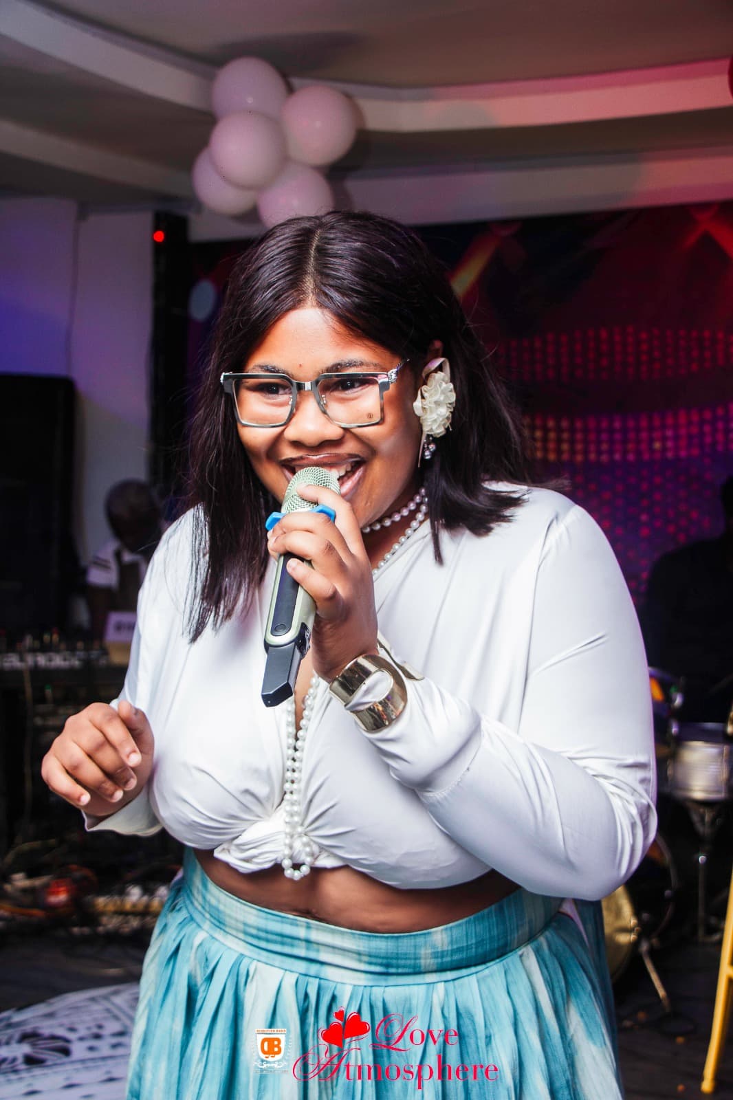 Smiling woman in glasses and white top singing into a microphone at a festive event.