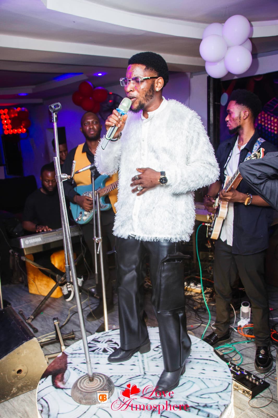 A male singer in a white fuzzy shirt performs on stage with a live band.