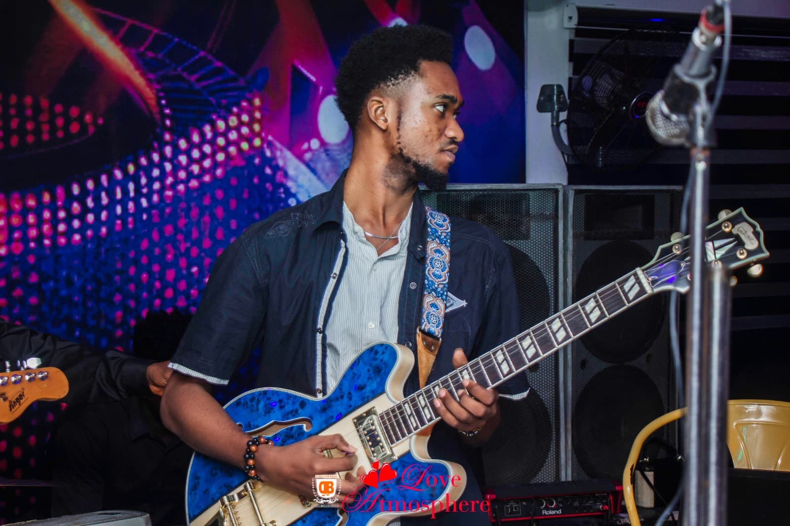 Bearded young man playing a blue electric guitar on stage with colorful background lighting.